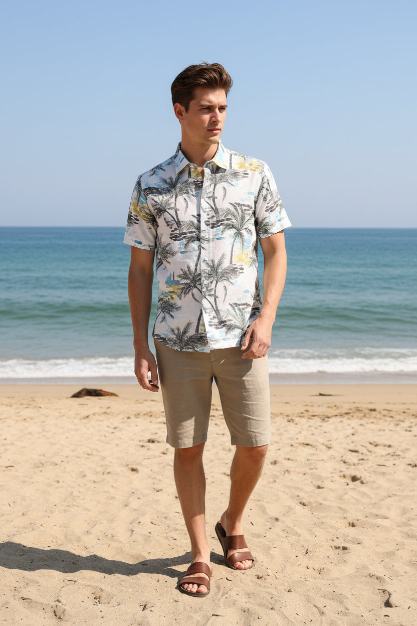 A folded long-sleeve button-up shirt by Hechter Paris featuring a tropical beach print with palm trees, blue water, and yellow accents on a white base, laid flat on a dark wood surface.