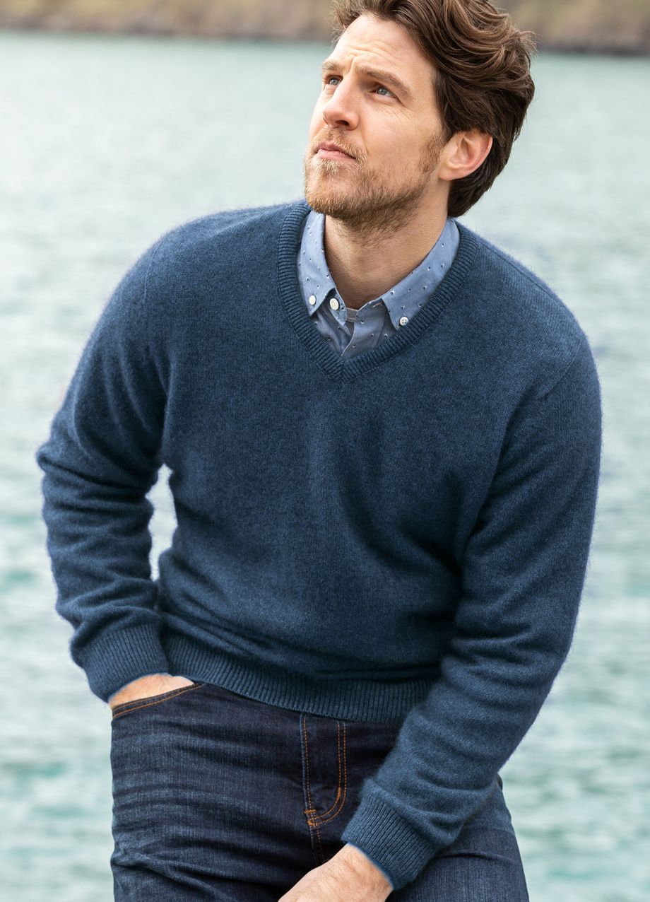 Man in navy V-neck sweater layered over button-down shirt, paired with dark jeans, by water