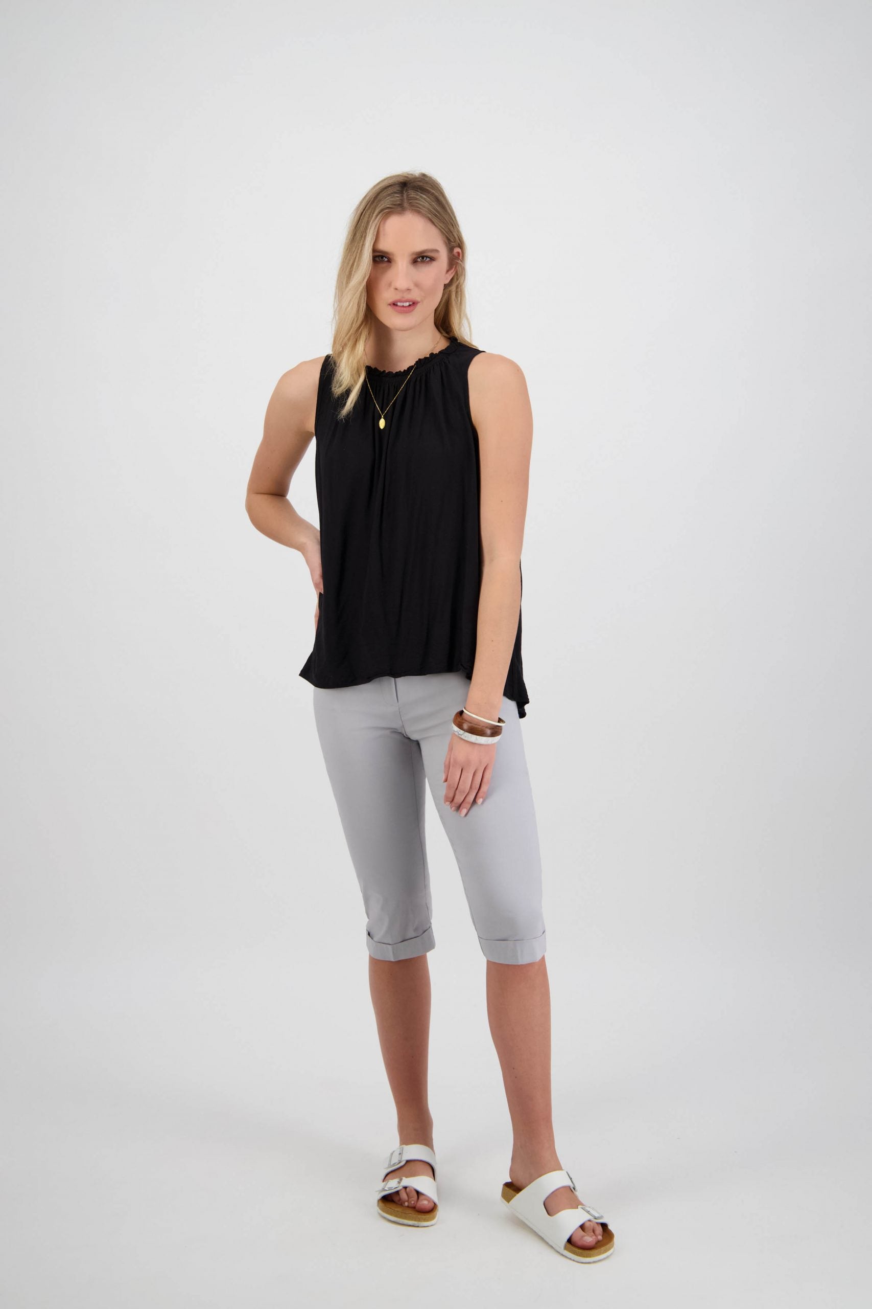 Woman wearing a black sleeveless blouse, grey cropped trousers, and white sandals.
